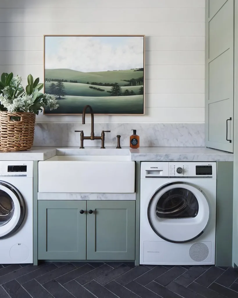 A modern farmhouse laundry room with a farmhouse sink, green cabinets, a washing machine, dryer, basket of flowers, soap dispenser, and a framed landscape painting above the sink.