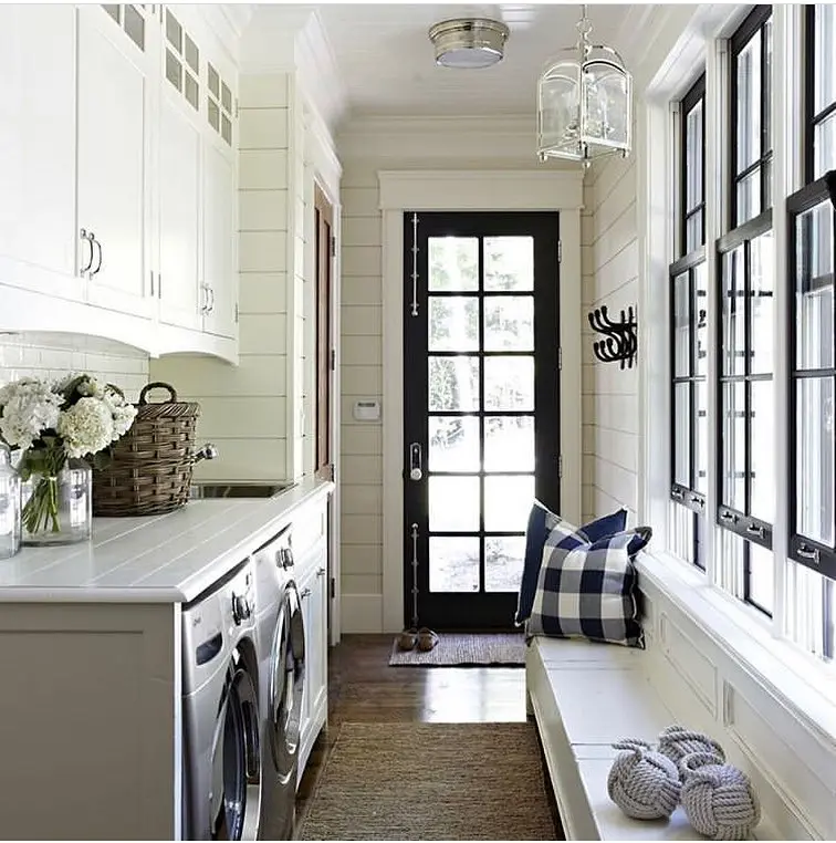 A bright modern farmhouse laundry mudroom features white cabinets, a washer and dryer, a bench with a plaid pillow, large windows, and a glass door leading outside.