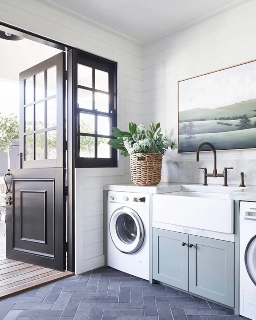 Modern farmhouse laundry room with a farmhouse sink, washing machine, blue cabinets, a basket with a plant, landscape painting, and a Dutch door open to the outside.