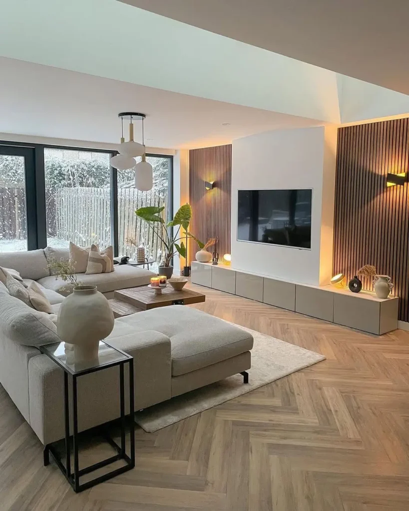 Modern living room with a light sectional sofa, wooden coffee table, wall-mounted TV, large windows, and wood panel accents. A stylish plant adds a touch of greenery while snow is visible outside through the windows.