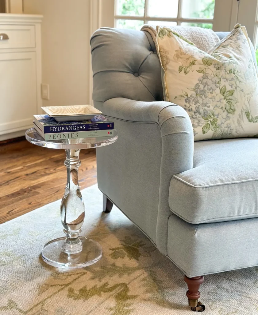 A light blue armchair with a floral pillow sits next to a small glass side table holding two flower books in a bright, cozy living room.