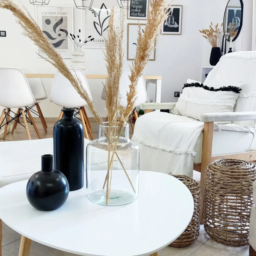 A modern living room with white furniture, a round white coffee table, and a clear vase holding tall dried grass. Black decorative items add contrast while artworks are displayed on the wall in the background.
