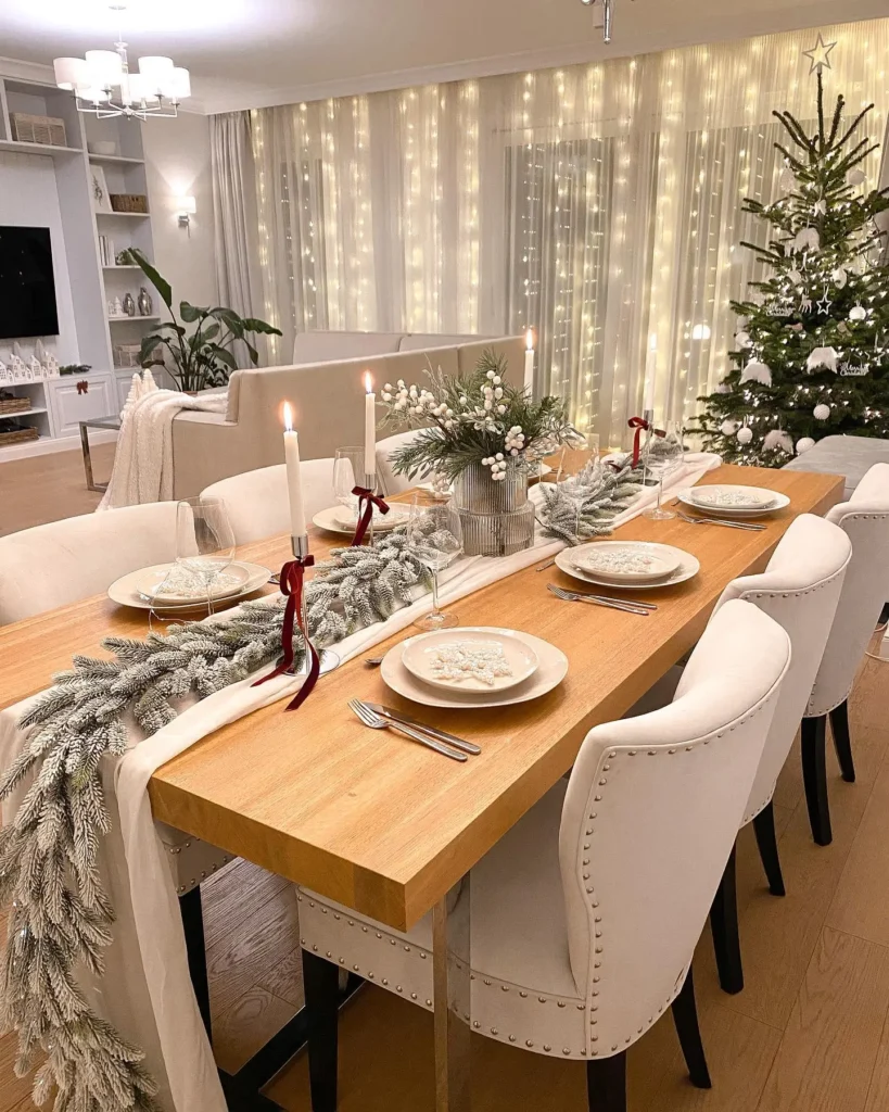 Elegant Christmas dining room with a wooden table decorated with a frosted garland, white candles, festive greenery centerpiece, and neutral upholstered chairs, set against warm string lights and a decorated Christmas tree.