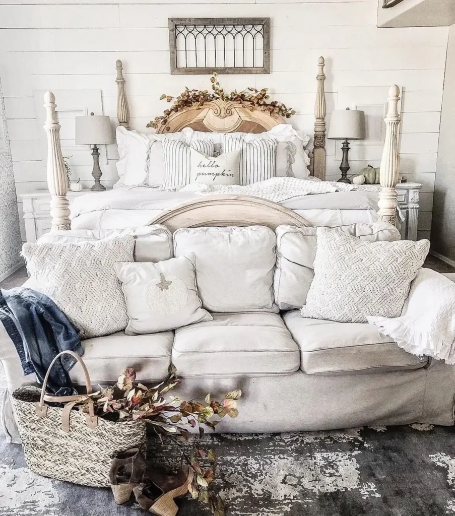 A white sofa with textured pillows sits at the foot of a bed with a wooden headboard, flanked by two bedside tables; a woven basket with foliage and shoes on the rug adds to the cozy fall farmhouse vibe.