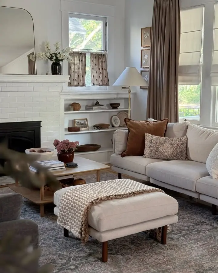 A cozy, earthy living room with a beige sectional sofa, patterned pillows, a knit throw on an ottoman, a wooden coffee table, built-in shelves, and a white brick fireplace brings organic modern warmth—perfect for fall.