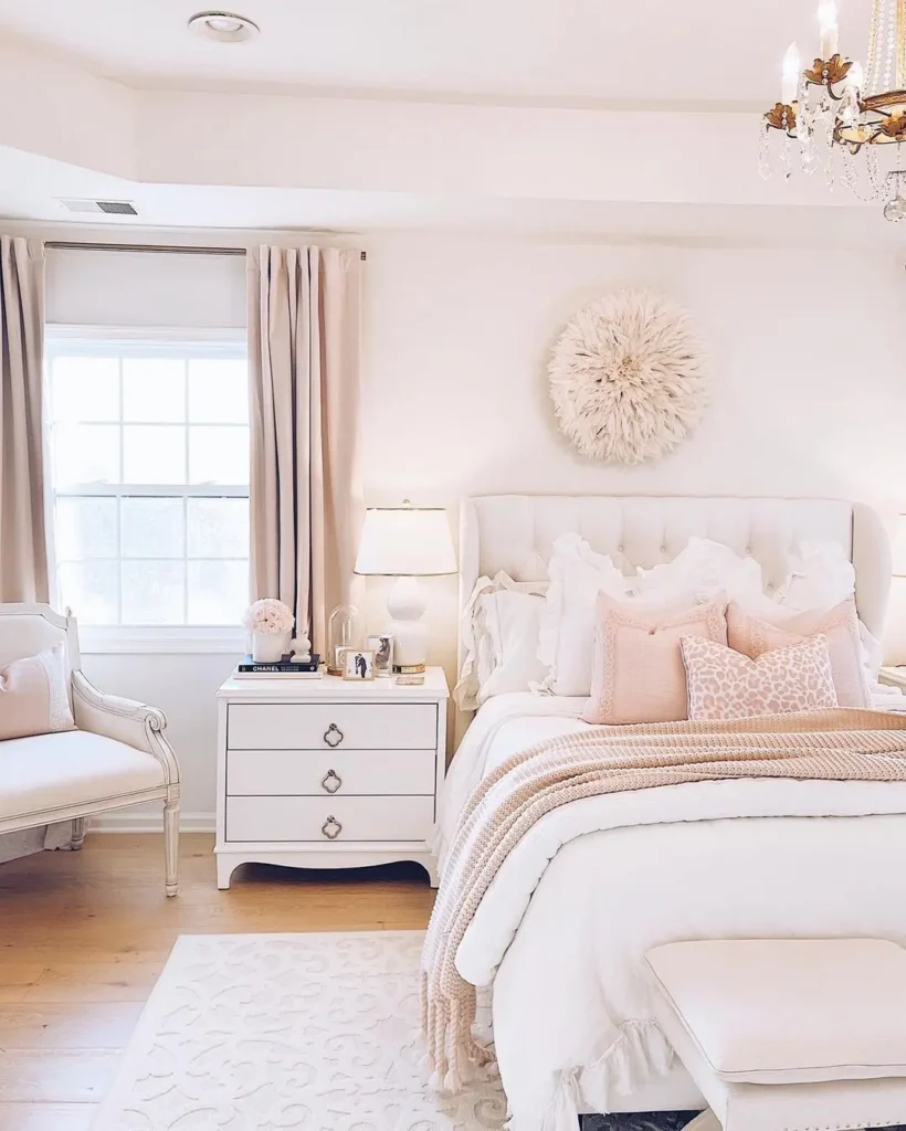 A bright bedroom with a light pink bed, pink pillows, a nightstand with a lamp, a window with beige curtains, and a bench by the wall. Neutral tones and soft textures throughout.