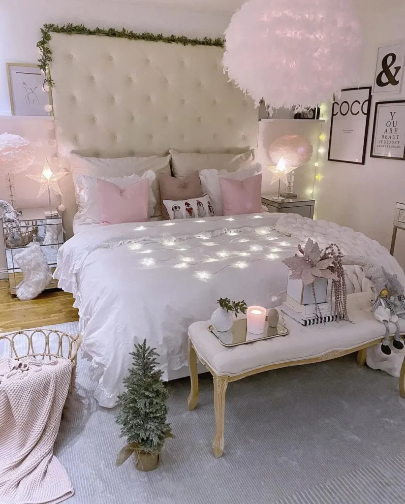 A bedroom with a large tufted headboard, white bedding, pink pillows, string lights, framed wall art, a small bench, candles, wrapped gifts, and miniature Christmas trees.