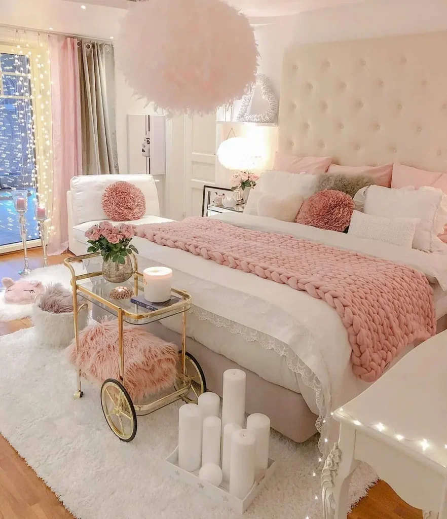 A pink-themed master bedroom with a large tufted headboard, pink and white bedding, plush pillows, a cozy knit throw, a gold cart with decor, candles, and soft lighting from pink fairy lights.