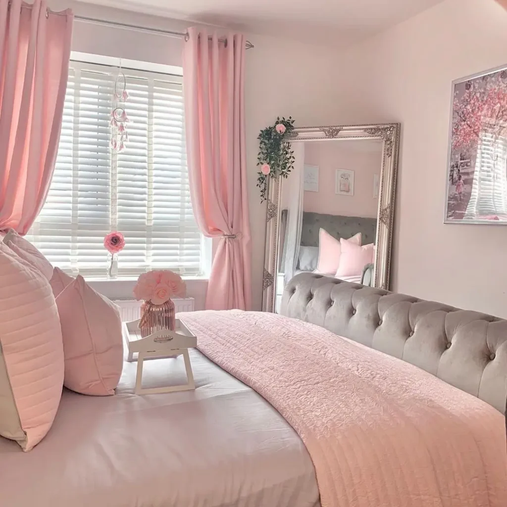 A neatly arranged bedroom with pink wall and curtains, bedding, and decor, a tufted gray headboard, a large mirror, and sunlight coming through white blinds.