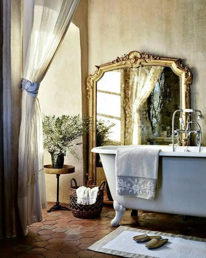 Elegant French country bathroom with a clawfoot tub, ornate gold-framed mirror, sheer curtains, potted plant, wicker basket, and a white rug with slippers on a tiled floor.