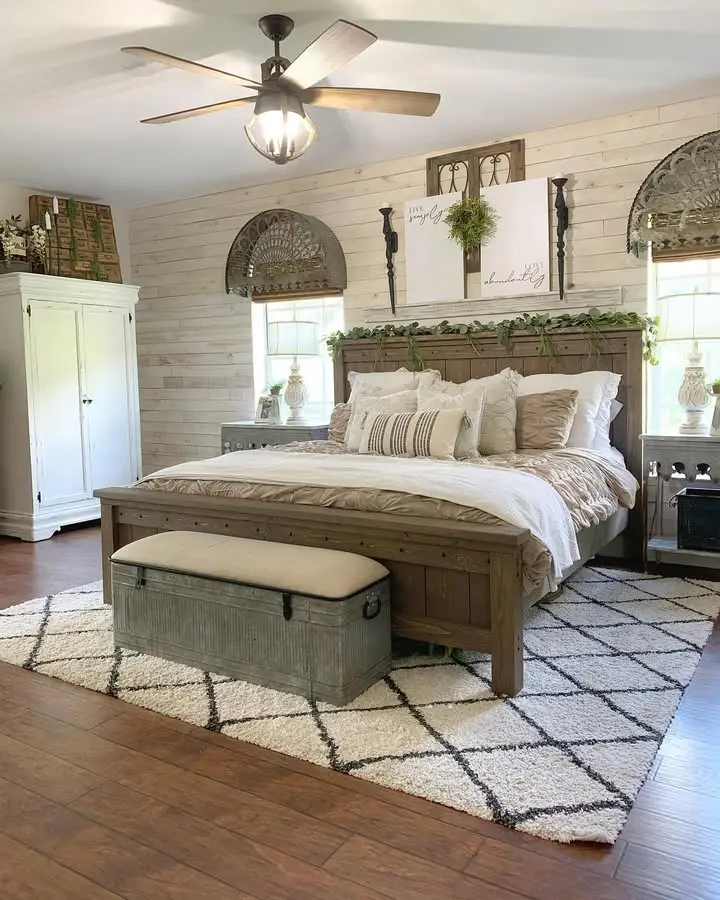 A bedroom with a wooden bed, white bedding, a gray metal bench at the foot, white wardrobe, rug, ceiling fan, and farmhouse-style decor on shiplap walls featuring ways to decorate with affordable rustic bedroom furniture.