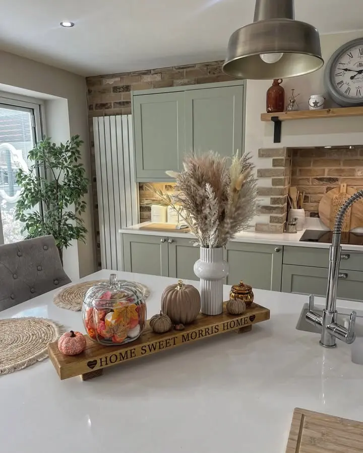 A modern kitchen with rustic sage green cabinets, a white countertop, a tray with autumn decor, and a vase of dried grasses on the island. A sign reads "Home Sweet Morris Home.