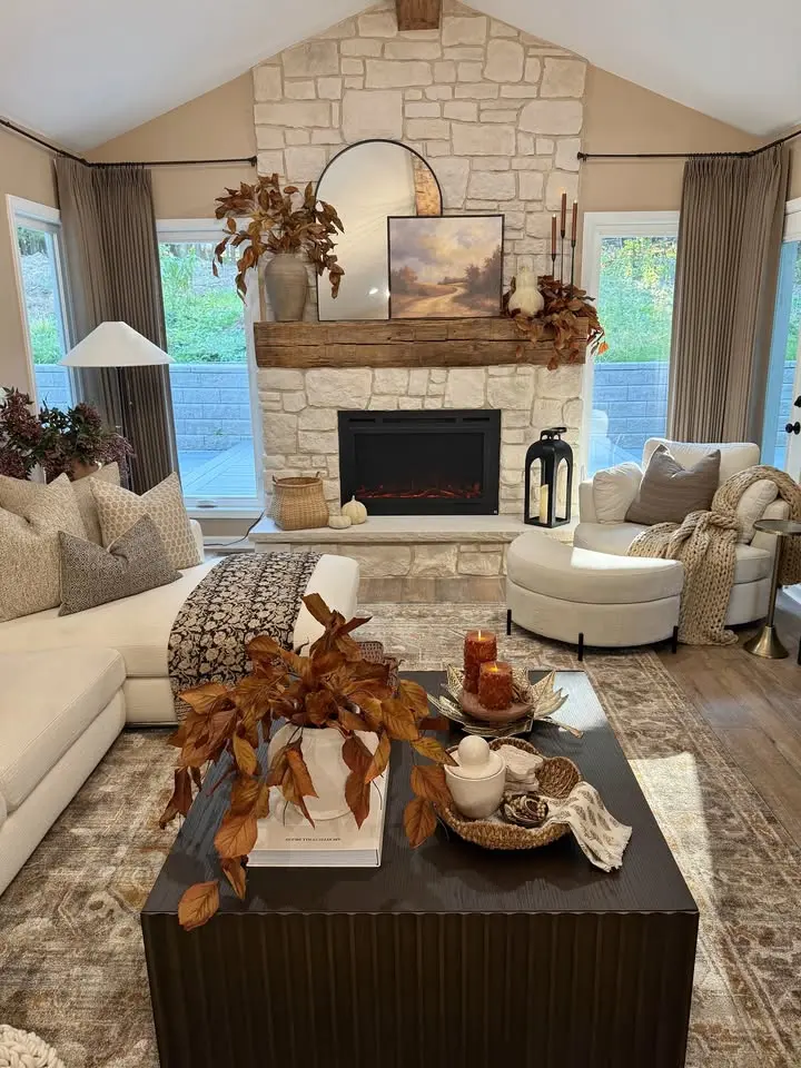 Modern living room with a stone fireplace, neutral furniture, autumn-themed decor, and large windows with drapes. The dark coffee table displays fall leaves and candles, perfect fall decor that transitions to winter seamlessly.
