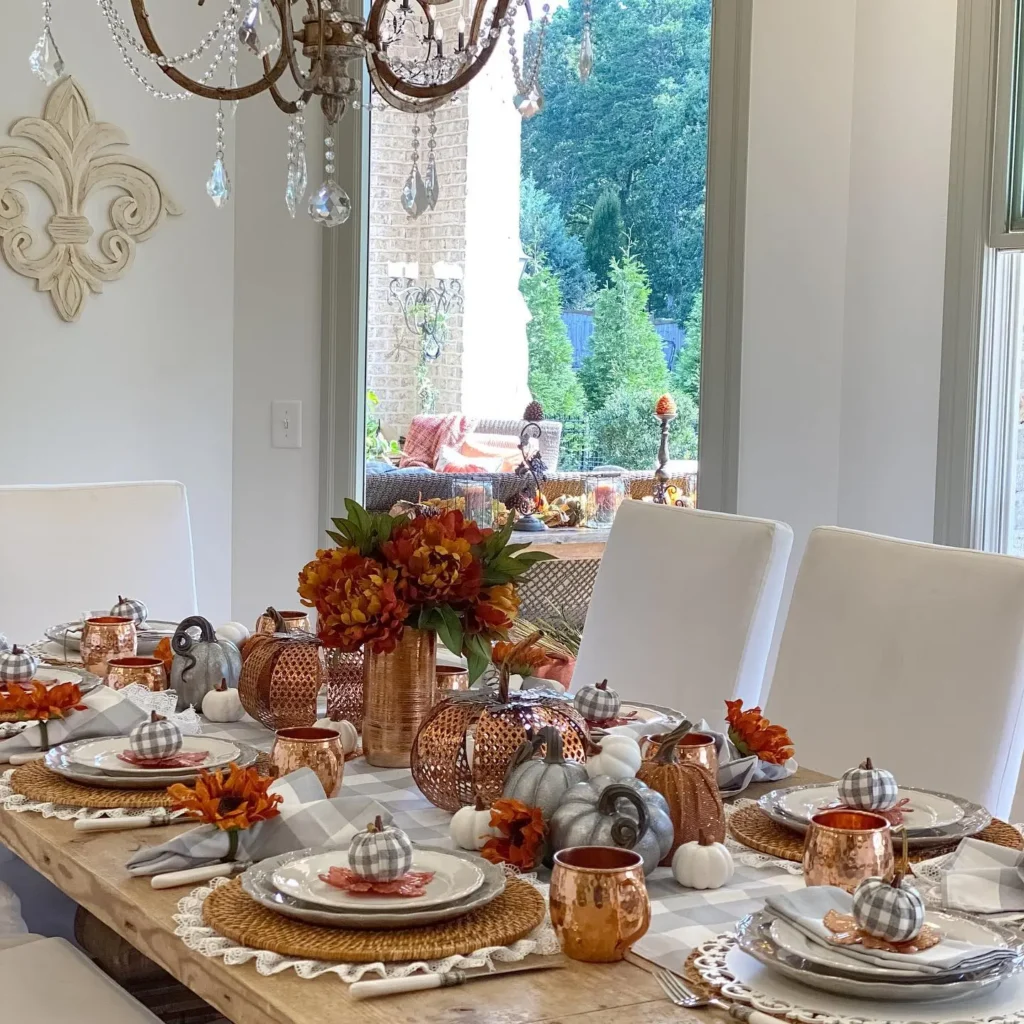 A dining table is set for fall with white plates, copper mugs, plaid napkins, a rustic centerpiece of small decorative pumpkins and orange flowers, and woven placemats in a bright, elegant room.