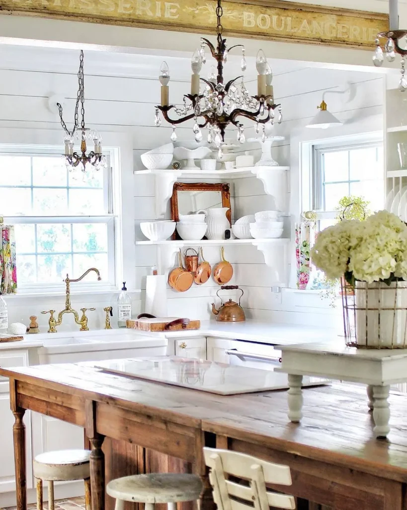 A bright kitchen with white cabinetry, rustic French country open shelves displaying dishes, a wooden island, brass fixtures, chandeliers, and a vase of white hydrangeas on the counter.
