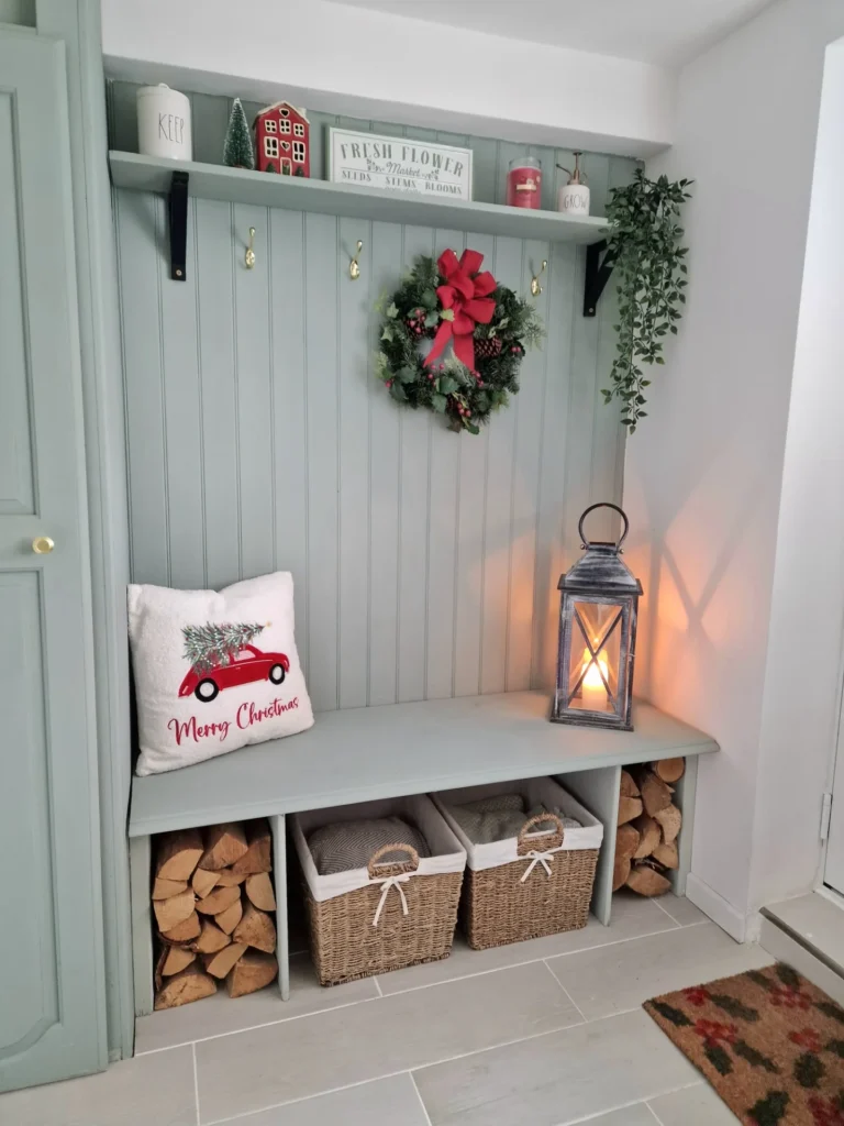 A cozy christmas mudroom features a hallway bench with a wreath, lantern, pillow, baskets, stacked firewood, and festive accents amid lush greenery.