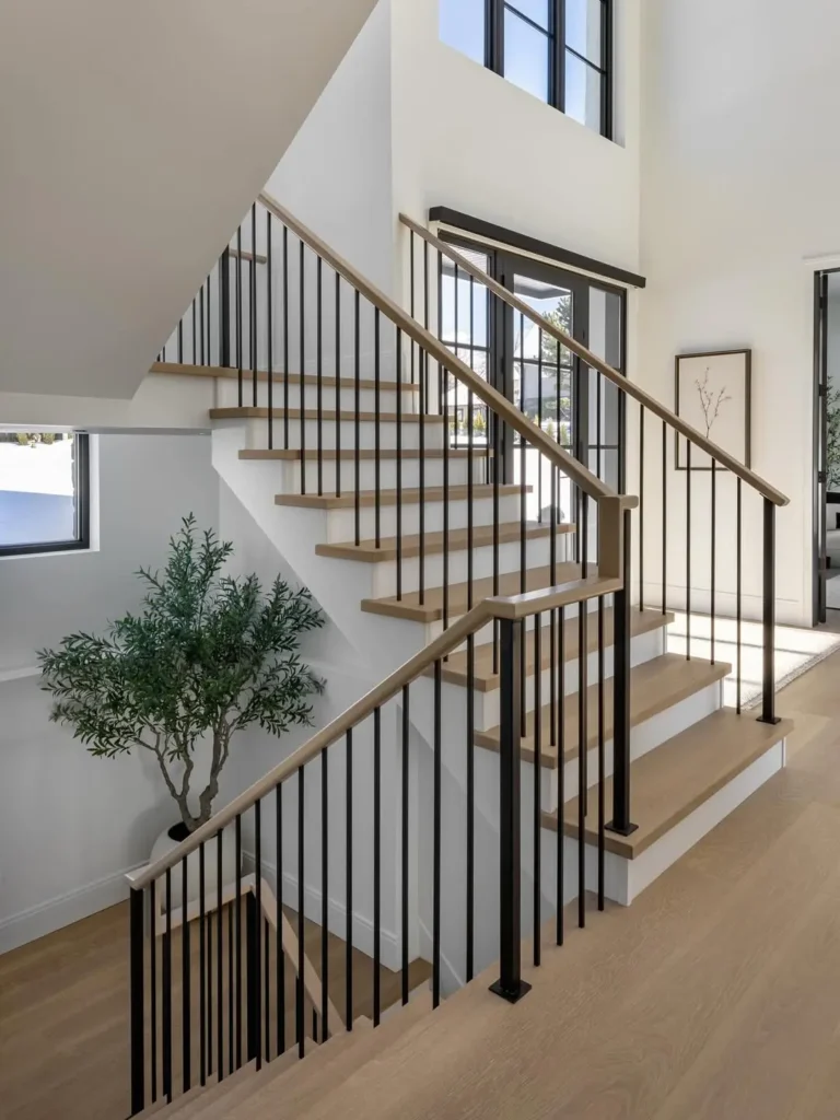 Modern indoor staircase with light wood steps, black metal railings, white walls, large windows, and a potted plant at the base.