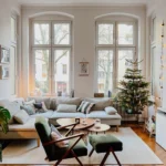 A cozy small living room with large windows, a decorated Christmas tree, neutral-toned furniture, wooden floors, and string lights along the wall, ready for Christmas celebration.