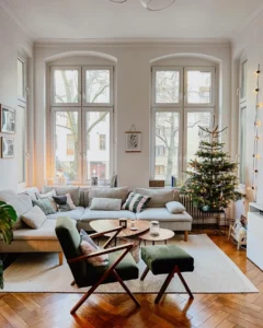 A cozy small living room with large windows, a decorated Christmas tree, neutral-toned furniture, wooden floors, and string lights along the wall, ready for Christmas celebration.