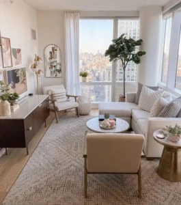 Bright, modern small living room with large windows, neutral-toned furniture, abstract wall art, a patterned rug, and cityscape views outside.