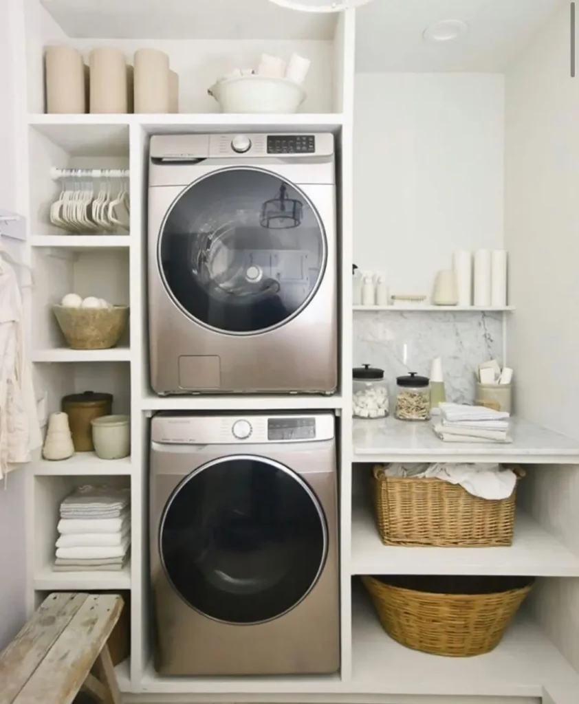 Modern small laundry room with stacked washer and dryer, open shelves holding laundry supplies, baskets, jars, and neatly folded towels in a neutral color palette.