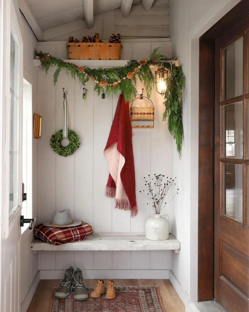 Cozy Christmas mudroom decorated with a natural evergreen garland, dried orange slices, woven baskets, a red throw blanket, a small boxwood wreath, winter boots, and a rustic bench styled with seasonal accents.