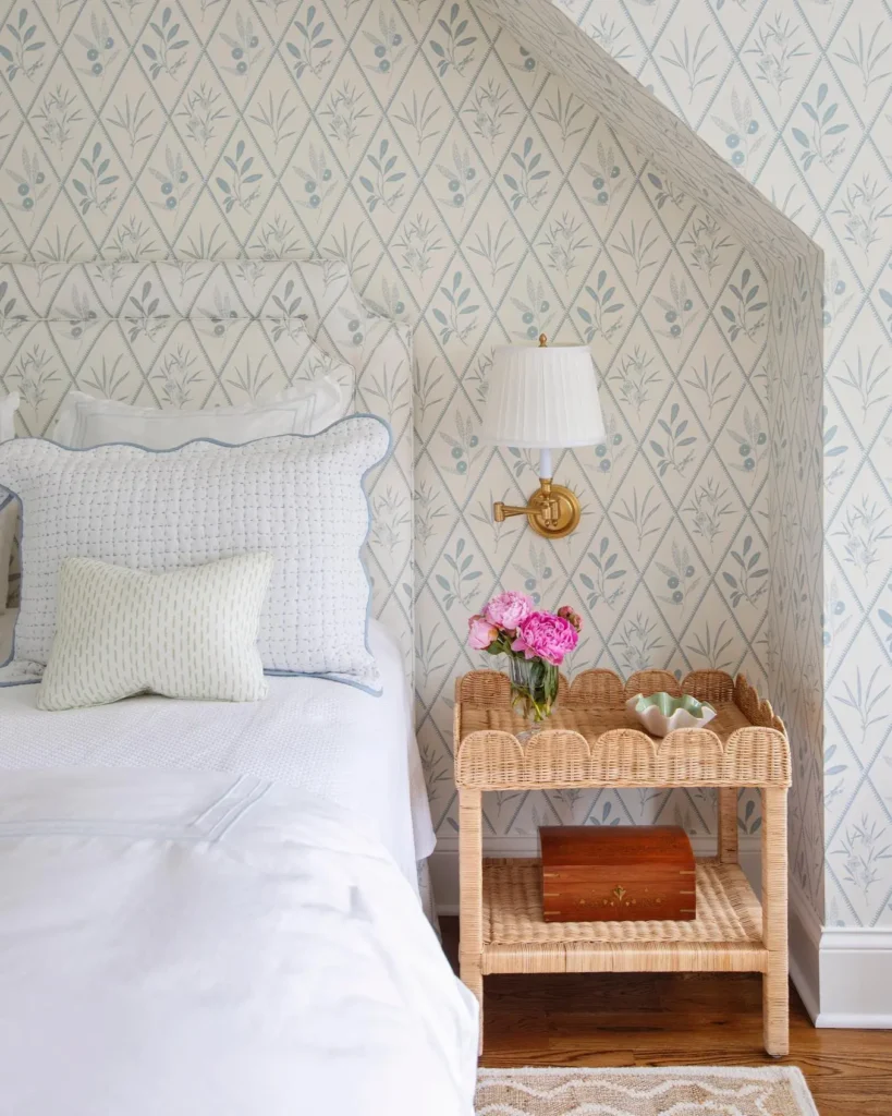 A neatly made bed with white bedding sits next to a wicker small side table holding pink flowers and a lamp, set against wallpaper with a blue floral pattern.