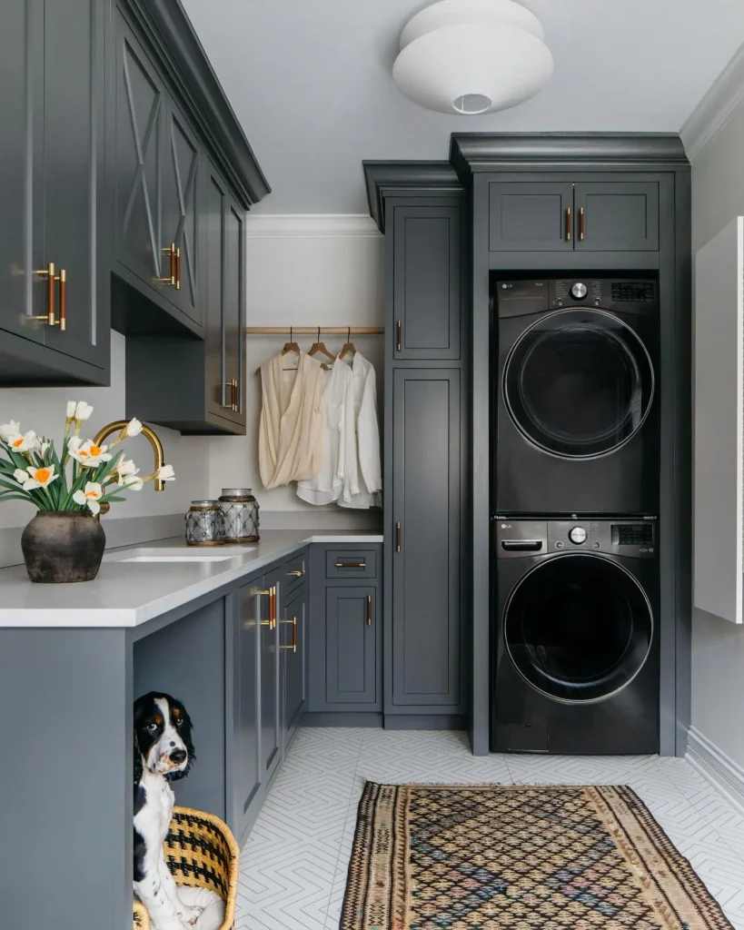 Modern laundry-mudroom with dark gray cabinets, stacked black washer and dryer, hanging shirts, a patterned rug, vase of flowers, and a dog sitting in a basket under the counter.