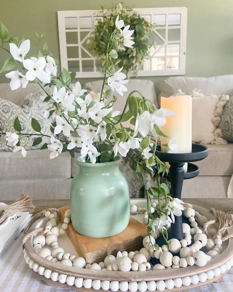 A cozy living room features a beige sofa with pillows, a green vase with white flowers on a decorative tray on the coffee table, and a wreath above a window frame on the wall, perfect elements for seamless seasonal transitions.