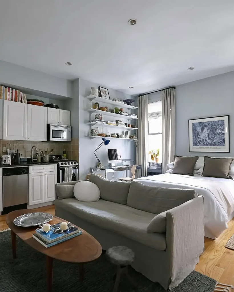A stylish studio apartment featuring a kitchen, open shelves, a desk by the window, a cozy bed with pillows, and a gray sofa facing a wooden coffee table.