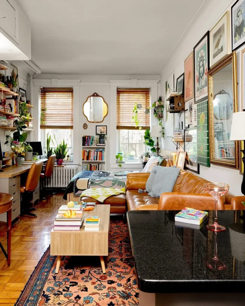 A cozy, well-lit studio apartment with vintage decor, featuring a bed, brown leather sofa, wall art, houseplants, bookshelves, and a desk by the window on wooden floors with a patterned rug.