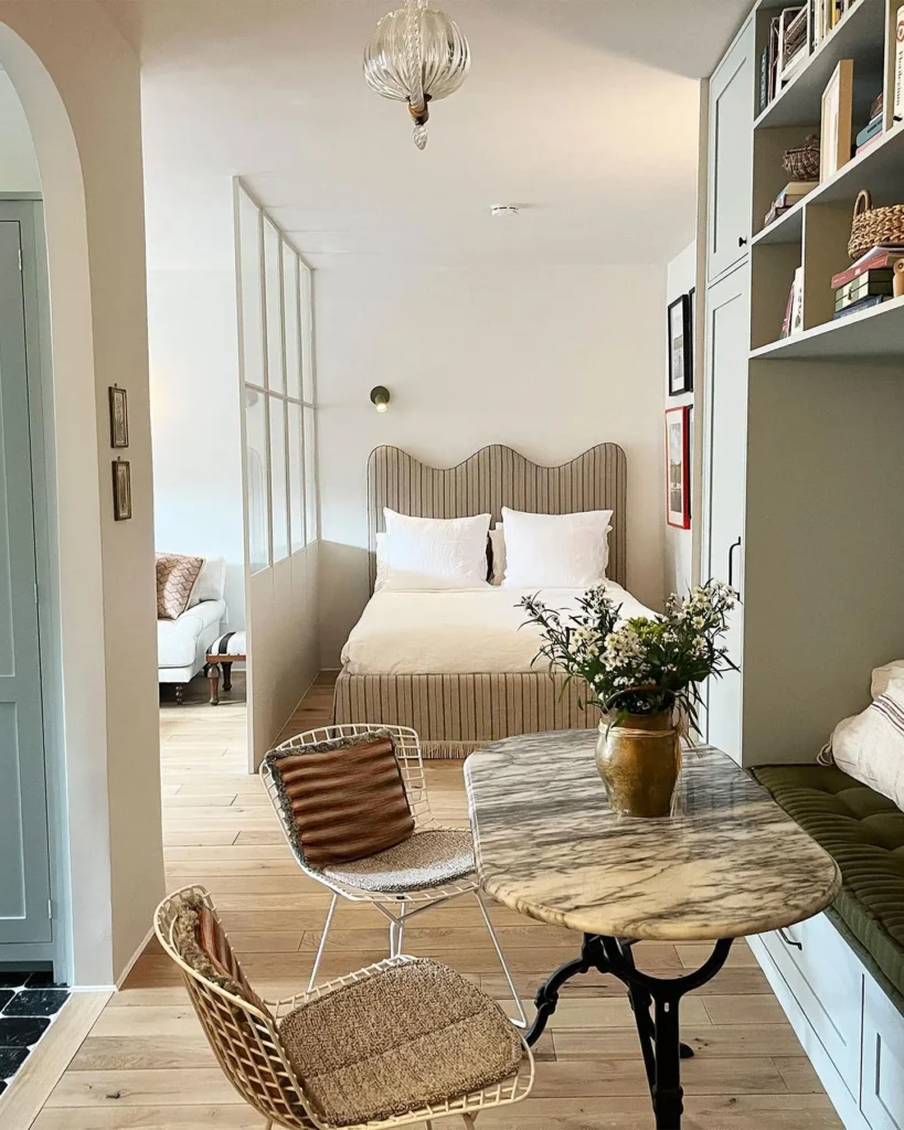 This cozy studio apartment features a bed with a striped headboard, a marble table with wicker chairs, built-in shelves, and a vase of flowers, plus a small lounge area in the background.