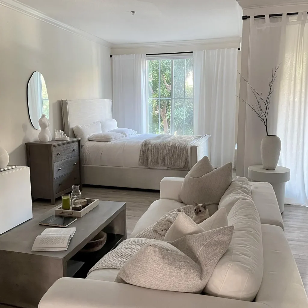 A modern, light-filled studio apartment bedroom features a white bed, a dresser with a mirror, a white sofa with pillows, a coffee table, and a large window with sheer curtains allowing natural light. A cat rests on the sofa.