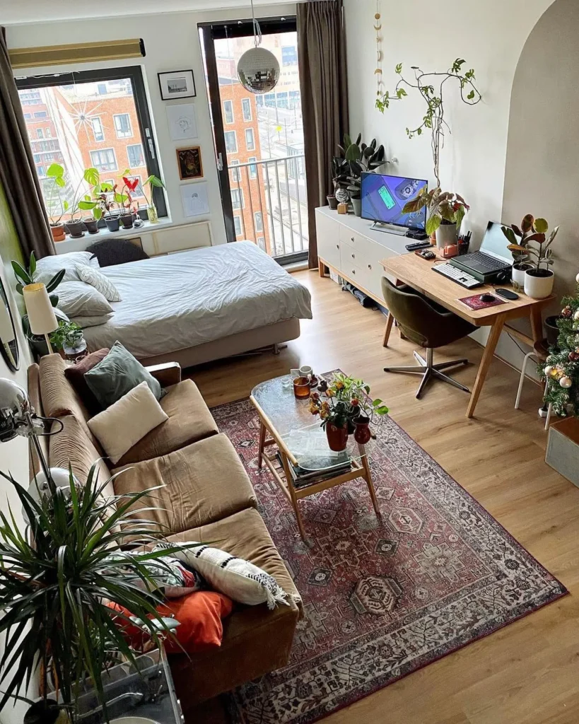 Modern studio apartment featuring a bed, sofa, coffee table, desk setup, lush houseplants, and large windows overlooking an urban view.