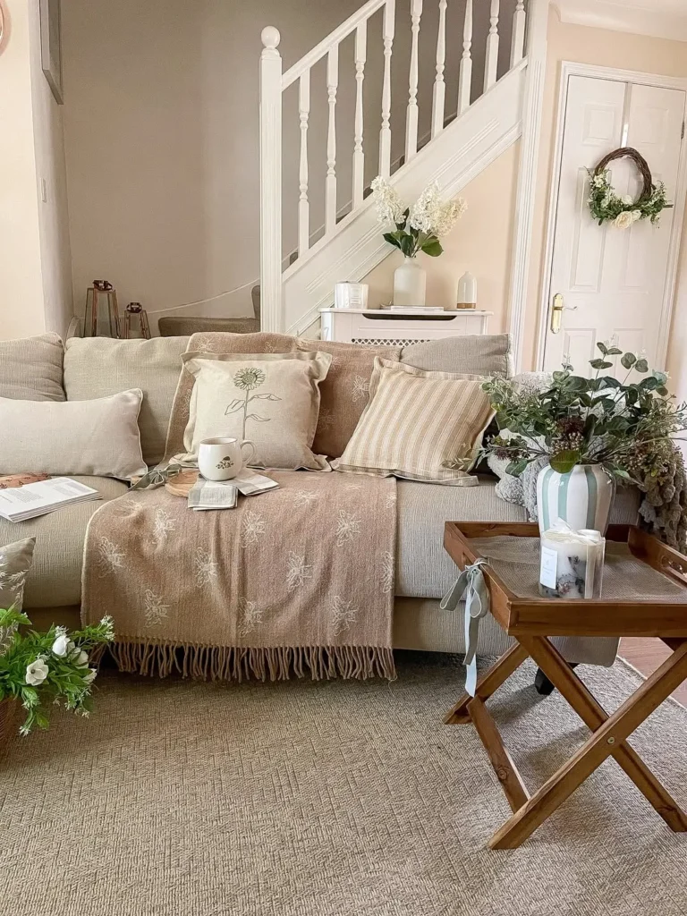 A beige living room with a sofa covered in neutral throw pillows and a throw blanket, a wooden tray table with plants, and a staircase in the background.