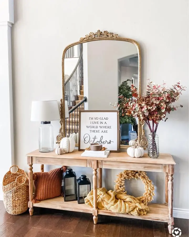 A wooden console table with pumpkins, vases of flowers, a lamp, lanterns, and a framed quote stands against a wall with a large ornate mirror above it.