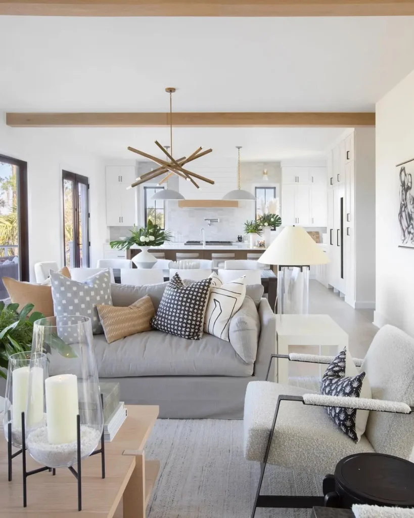 Modern open-concept living room and kitchen with neutral tones, a gray sofa with patterned pillows embracing textures and fabrics in throw pillow styling, armchair, candles, pendant light, and green plants.