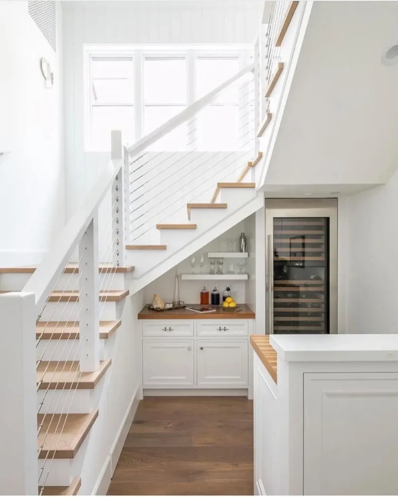 A small bar area with a countertop, cabinets, shelves, and a wine fridge is built under a white staircase in a bright, modern interior.