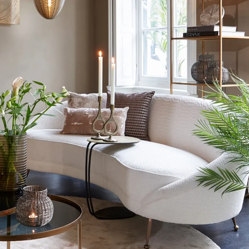 A modern living room with a white curved sofa, decorative pillows, a small side table featuring candles, green plants, and a round glass coffee table.