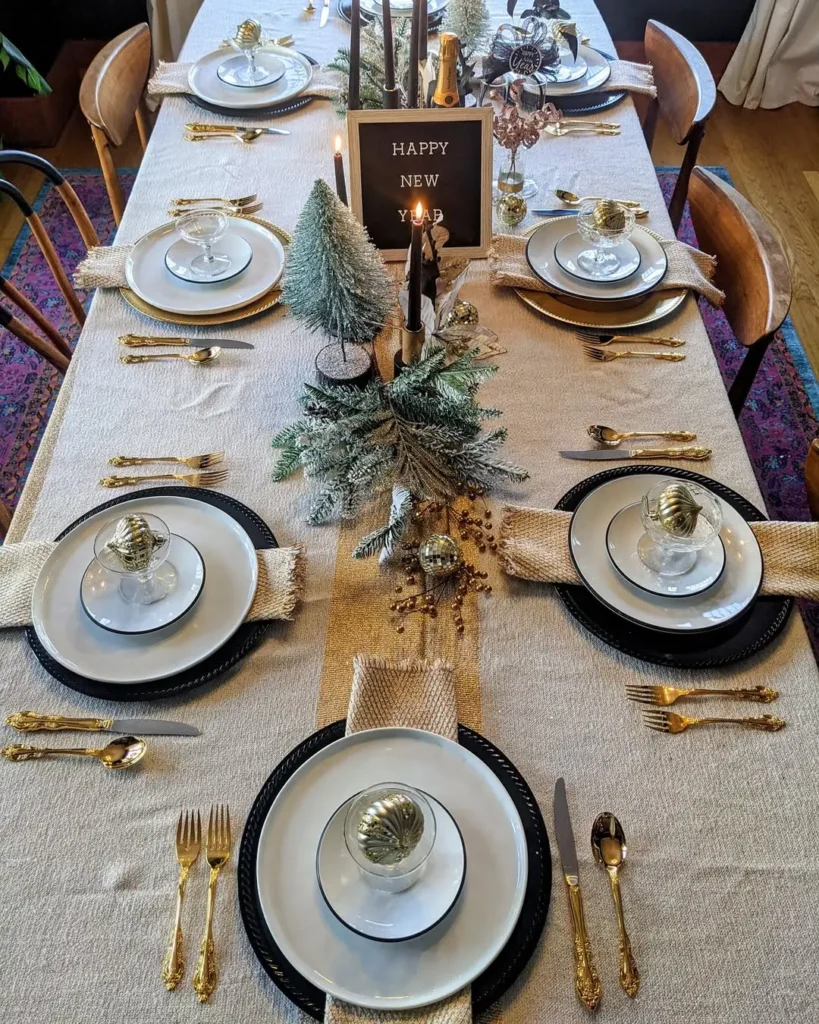 A rectangular dining table set for six with gold cutlery, white plates, festive winter table decorations, candles, and a "Happy New Year" sign as the centerpiece.