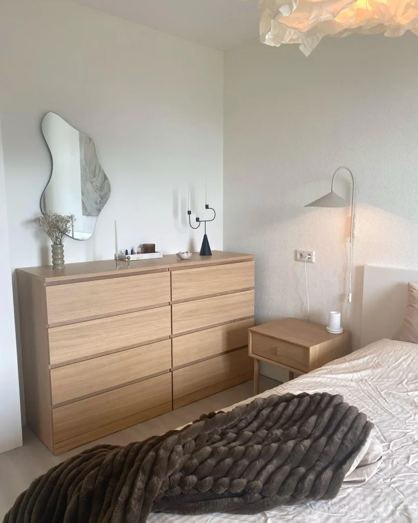 Minimalist scandinavian bedroom decor with a wooden dresser and nightstand, abstract mirror, white walls, and soft lighting. A brown textured blanket lies on the bed.