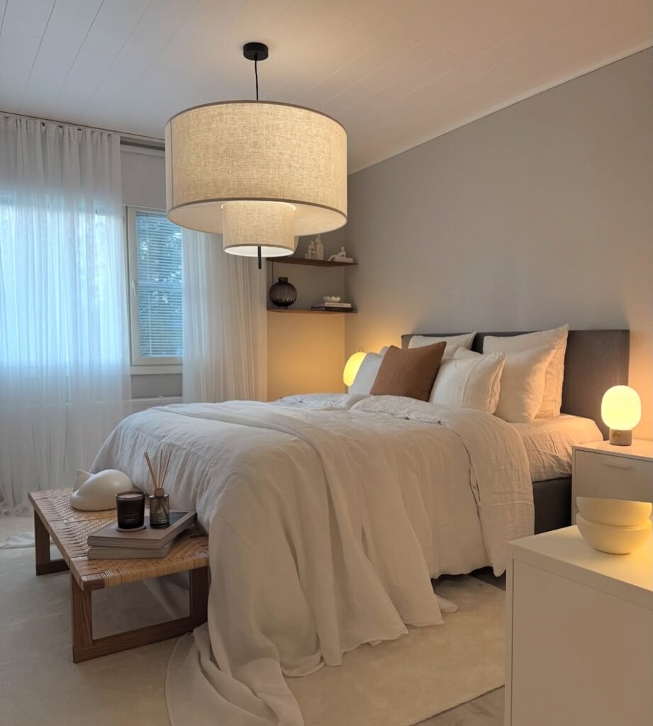 Modern bedroom with a neatly made bed, neutral bedding, Scandinavian bedroom decor touches, two bedside tables with lamps, a large pendant light, and a window with sheer curtains letting in daylight.