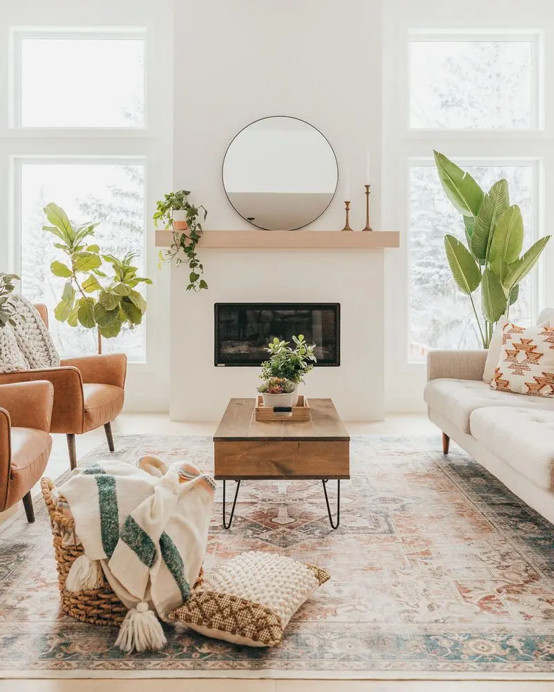 Bright bohemian living room with neutral tones, a round mirror above a fireplace, potted plants, a wooden coffee table, and chairs with patterned cushions and a throw in a woven basket.