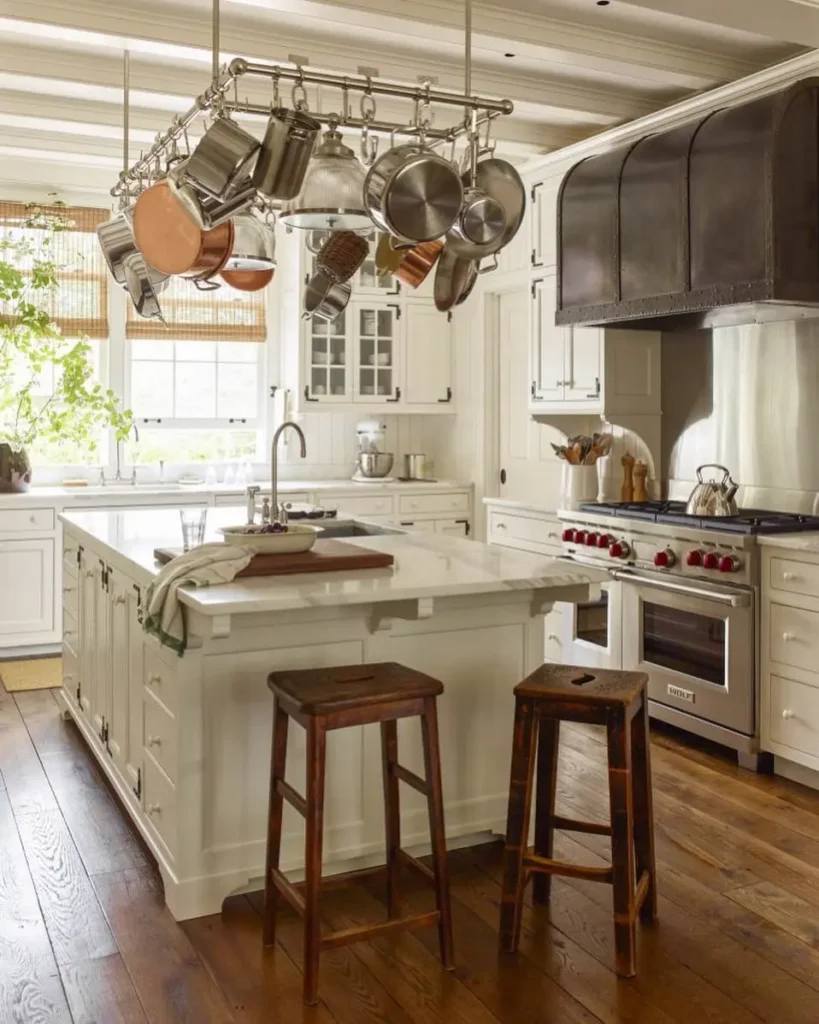 A bright kitchen with white cabinets, a central island with two wooden stools, a ceiling pot rack displaying hanging pots and pans, a large stove with a metallic hood, and warm wooden floors.