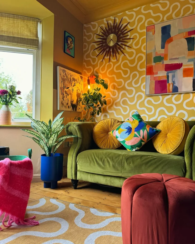 A living room with a green velvet sofa, patterned yellow walls, colorful artwork, houseplants, and eclectic accessories. Bright accent decor and a large window fill the space with natural light.