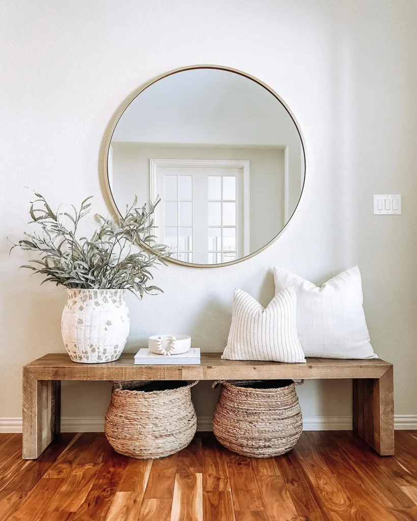 A wooden bench with two woven baskets, white pillows, and a large vase with greenery sits below a round wall mirror in a bright, minimalist entryway.
