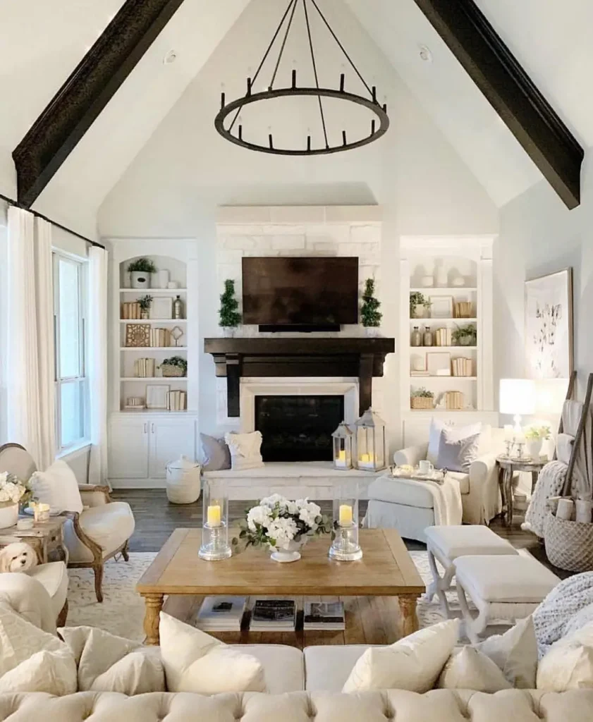 Bright farmhouse living room with vaulted ceiling, dark beams, chandelier, fireplace, built-in shelves, neutral furniture, and decorative accents including candles and flowers.