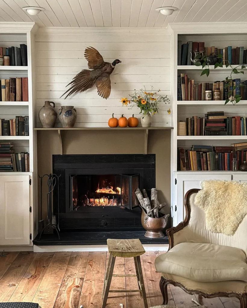A cozy farmhouse living room with a lit fireplace, built-in bookshelves, three pumpkins, vases, flowers, and a mounted pheasant above the mantel, with an armchair and wooden stool in front.