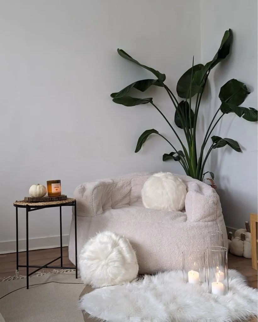 A cozy corner with a plush white chair, faux fur cushions, a small side table with a candle and pumpkins, a tall green plant, and lit candles on a white rug.