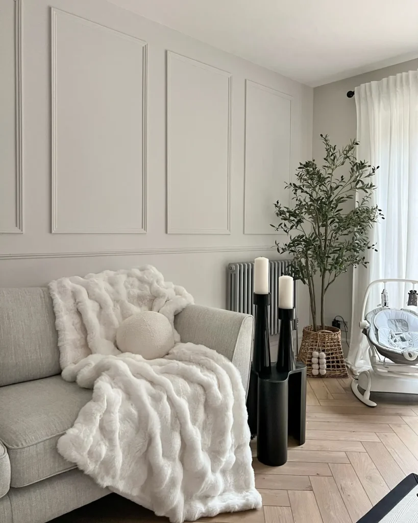 A modern living room with a light gray sofa, faux fur throw, round pillow, black candle holders, potted tree, woven basket, and a baby rocker near sheer curtains.